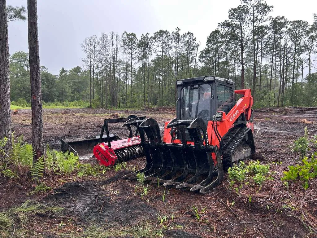 Kubota tracked skid steer with bucket and mulcher attachments at a wooded jobsite.